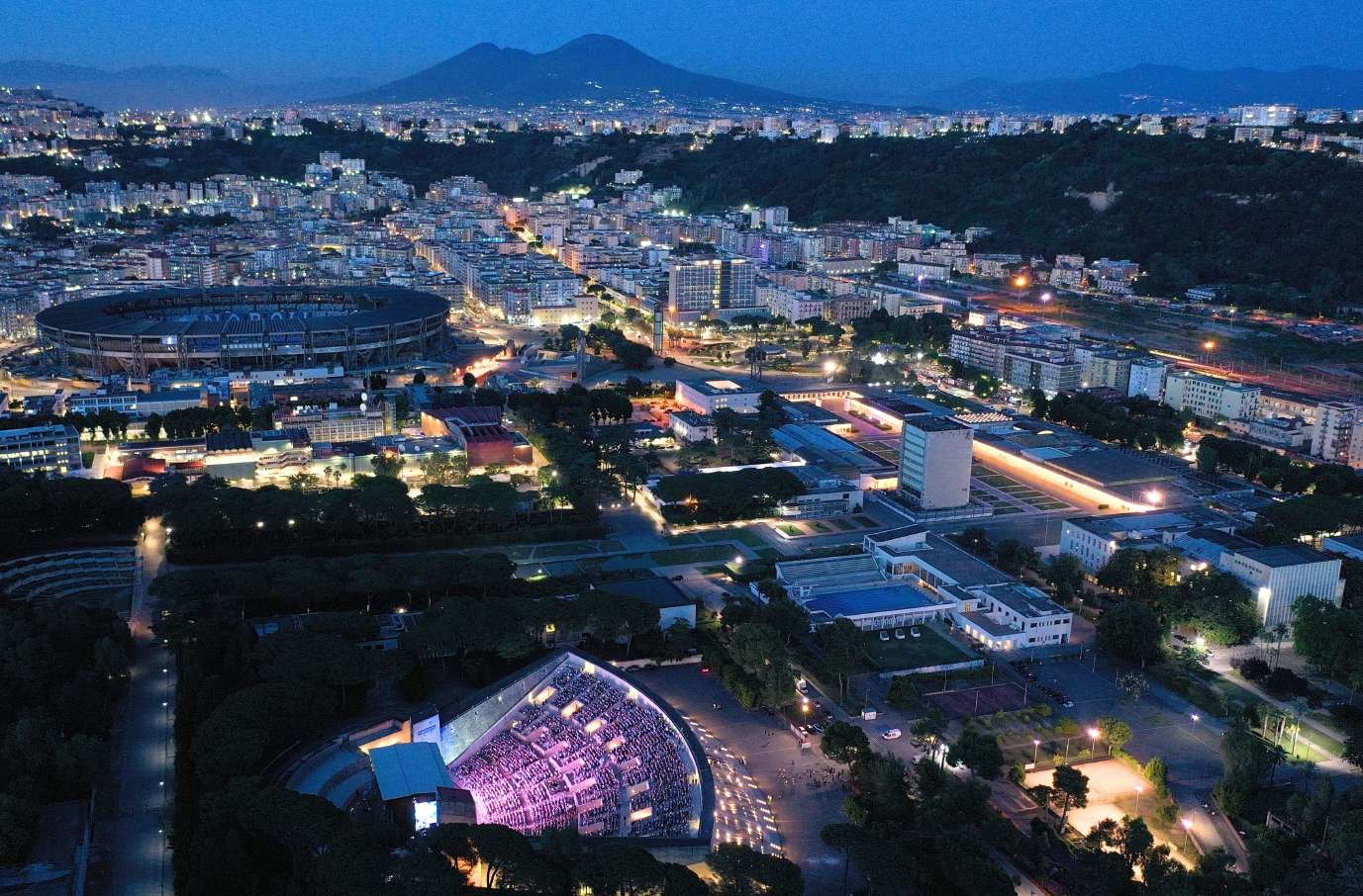 Fuorigrotta di sera vista dall'alto: l'Arena Flegrea illuminata in primo piano, lo stadio Maradona e il Vesuvio sullo sfondo